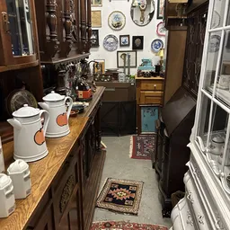 Smalle winkelgang met houten kasten, witte kannen met oranje accenten, borden aan de muur en diverse antieke objecten