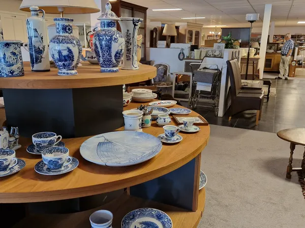 Winkelvloer met ronde houten toonbanken met blauwe servies en porselein uitgestald, op de achtergrond meer kasten en meubels