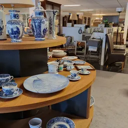 Winkelvloer met ronde houten toonbanken met blauwe servies en porselein uitgestald, op de achtergrond meer kasten en meubels