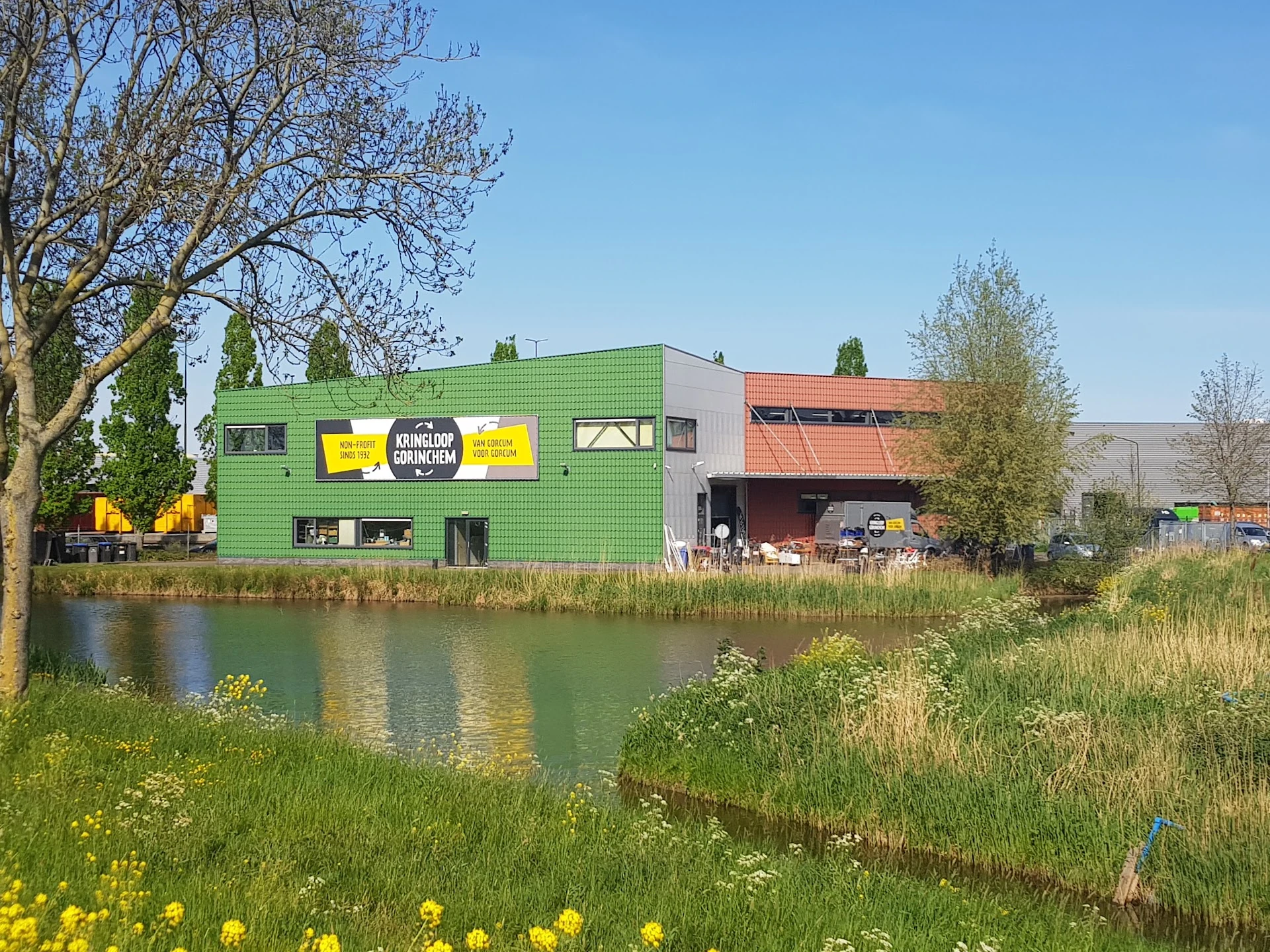 Groene gevel van kringloopwinkel aan water met gele reclameborden en bruine schuur ernaast