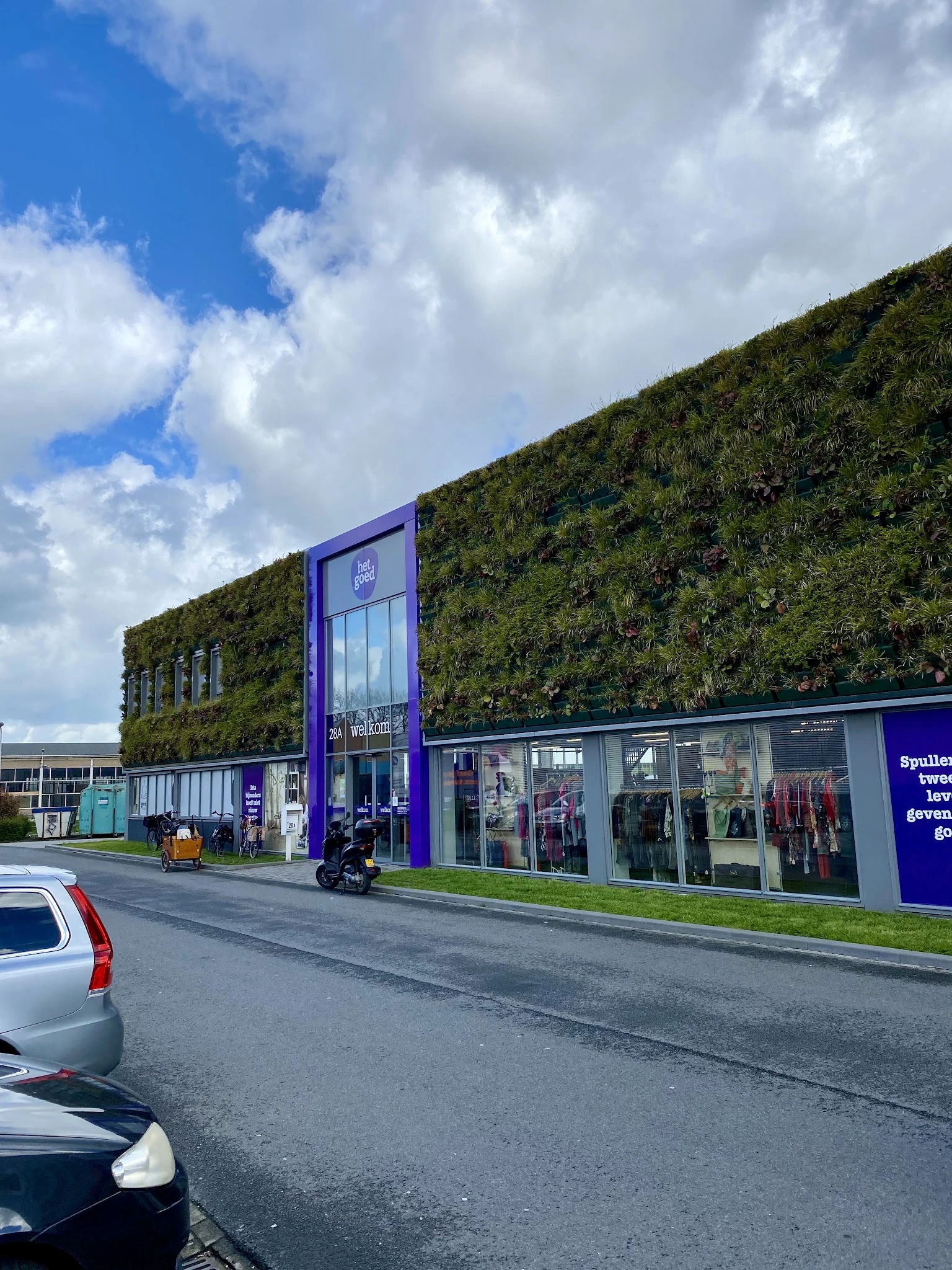 Moderne gevel van kringloopwinkel met groene plantenwand, paarse kozijnen en etalages met kleding