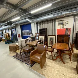 Diverse meubels in een kringloopwinkel, waaronder houten stoelen, tafels, een bruine fauteuil en kasten.