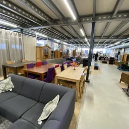 Ruime kringloopwinkel met meubels, tafels, stoelen en kasten uitgestald op een heldere vloer onder industriële verlichting