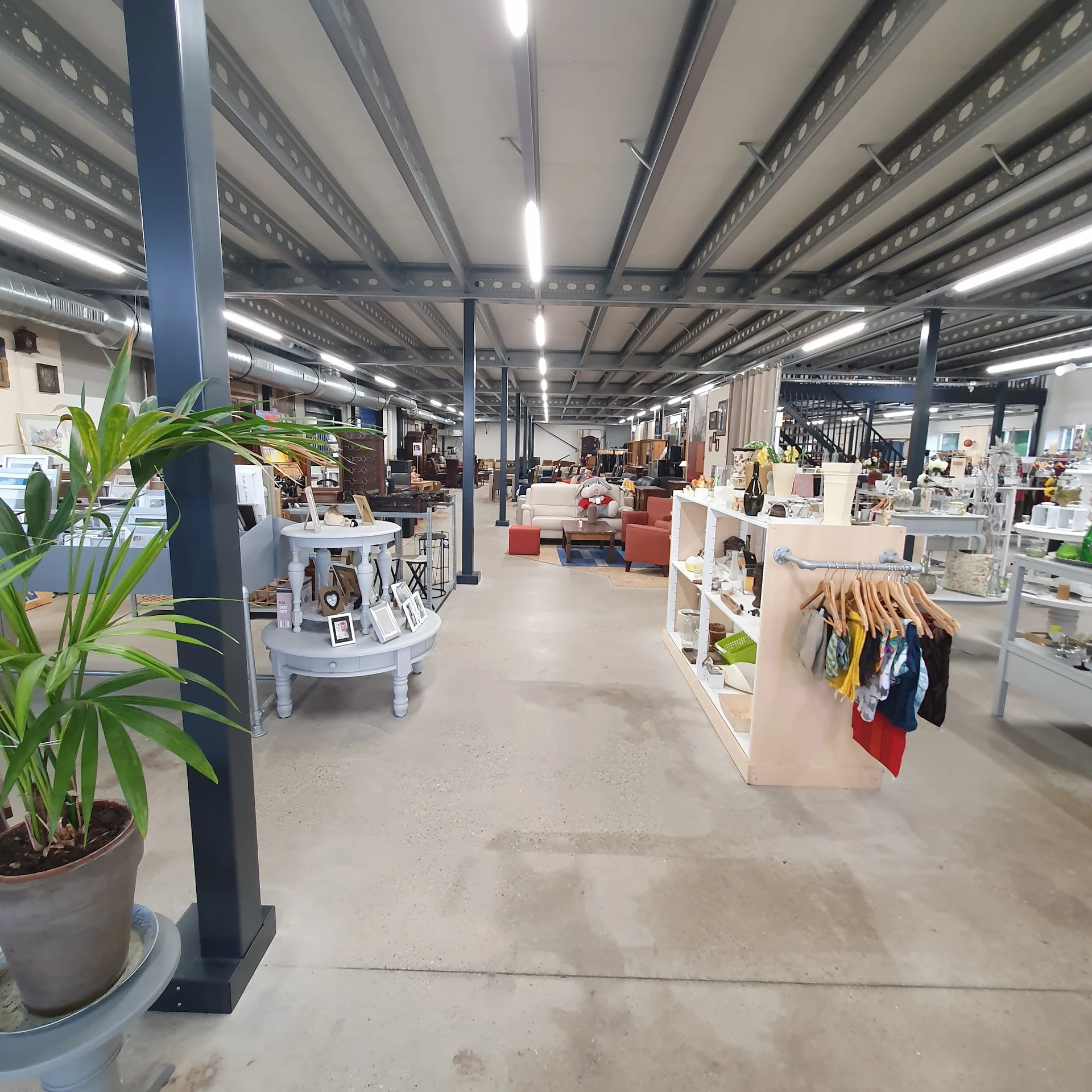 Ruim kringloopwinkelinterieur met stellingen, kledingrekken, meubels en decoratie verspreid over betonnen vloer onder industr