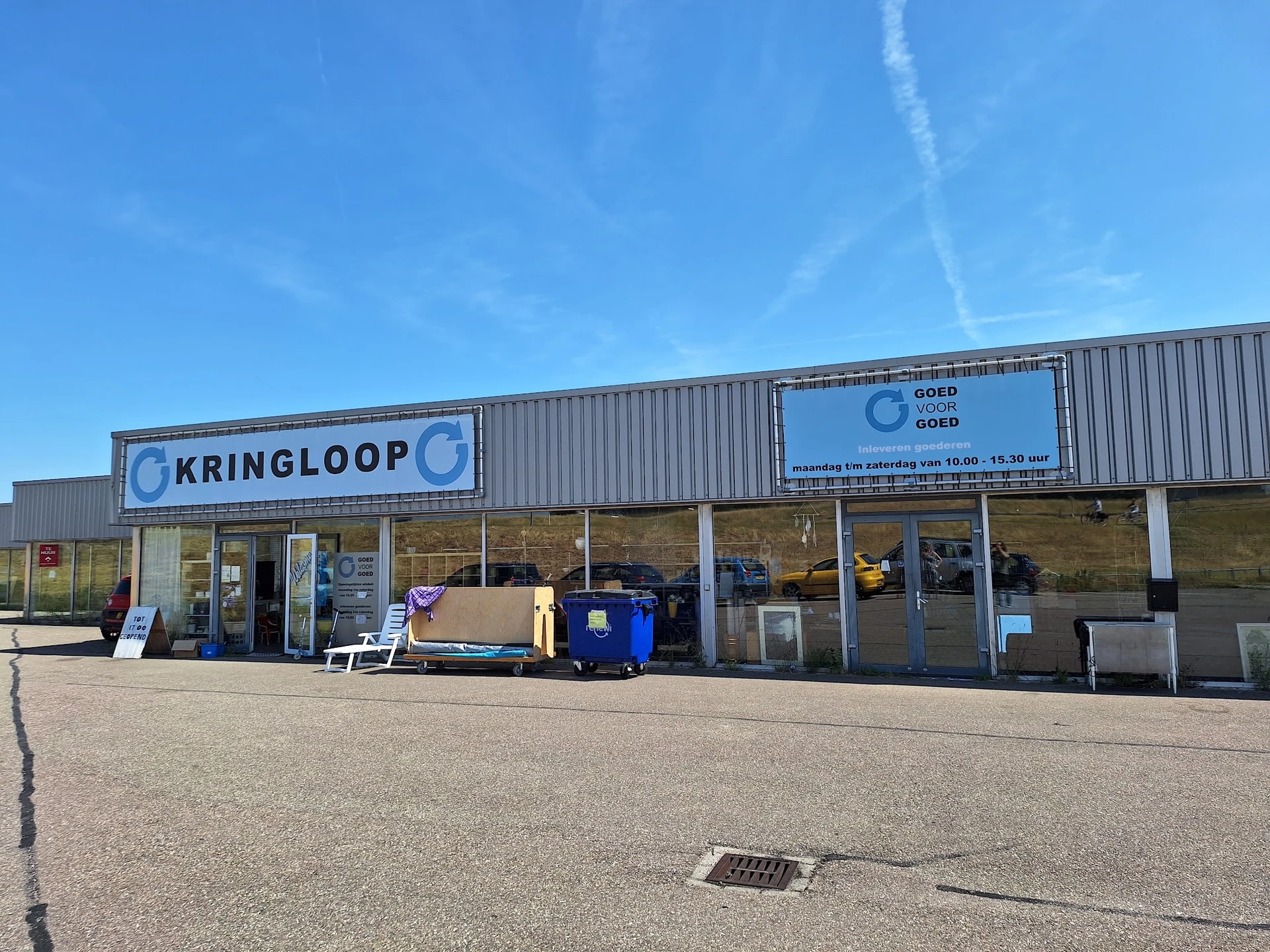 Gevel van kringloopwinkel met naam KRINGLOOP zichtbaar, enkele artikelen buiten uitgestald, heeldere dag