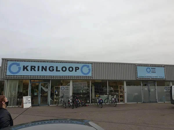 Voorgevel van kringloopwinkel met grote blauwe letters, ingang met glazen deuren en enkele fietsen ervoor