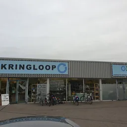 Voorgevel van kringloopwinkel met grote blauwe letters, ingang met glazen deuren en enkele fietsen ervoor