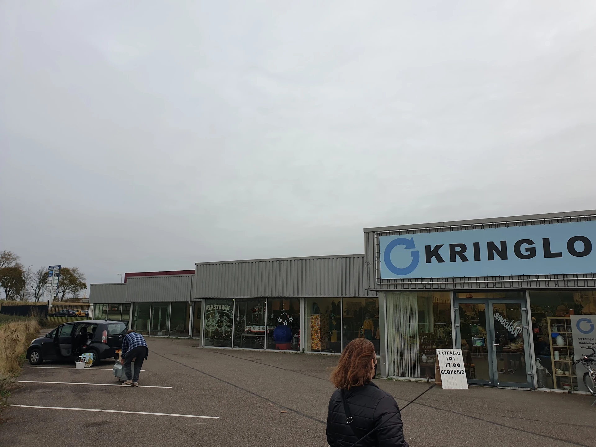Gevel van kringloopwinkel met naam 'KRRINGLO' op bord, parkeerplaats en voetganger zichtbaar