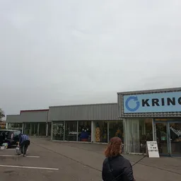 Gevel van kringloopwinkel met naam 'KRRINGLO' op bord, parkeerplaats en voetganger zichtbaar