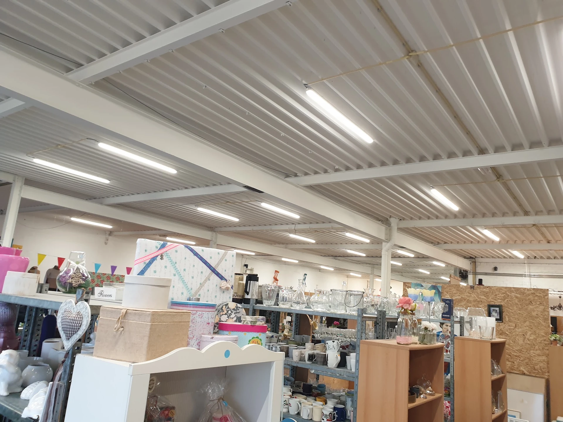 Binnenaanzicht van kringloopwinkel met stellingen, kasten en diverse gebruiksartikelen onder rioleringsdak met LED-verlichtin