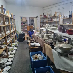 Binnenaanzicht van kringloopwinkel met houten stellingen vol servies, tafels met huishoudwaar en blauw sorteerkrat.