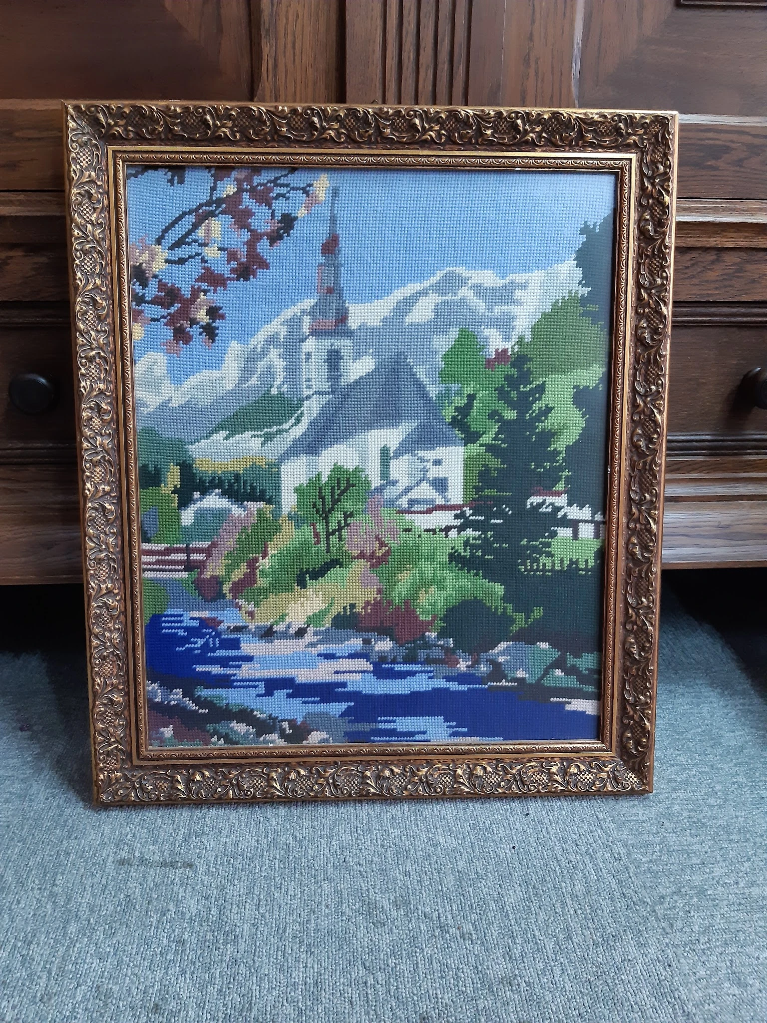 Ingelijste kruissteekwerk met berglandschap, kerk en rivier