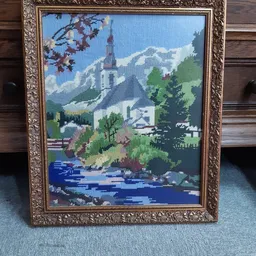 Ingelijste kruissteekwerk met berglandschap, kerk en rivier