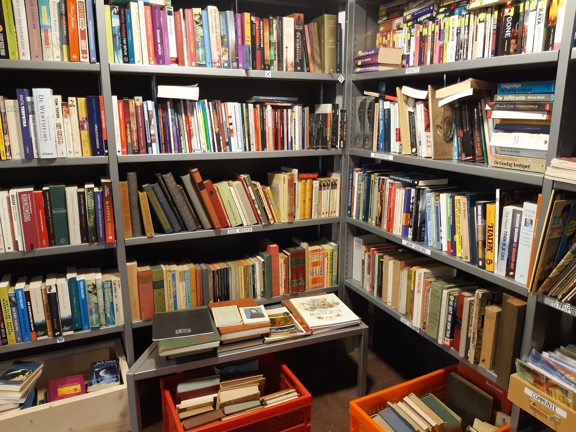 Boekenkasten vol met tweedehands boeken in verschillende genres en kleuren, met daaronder oranje bakken met extra boeken.