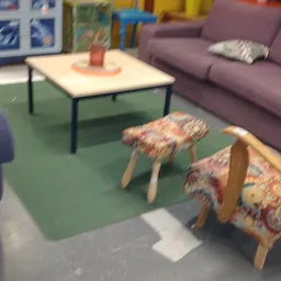 Zithoek met paarse bank, tafel en twee gevlochten krukken op groen tapijt