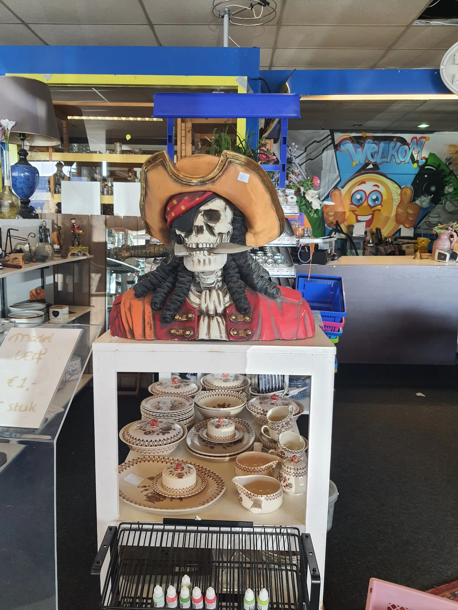 Uitgestalde decoratieve items en servies in de kringloopwinkel, inclusief een piratenskelletfiguur en aardewerk met patroon