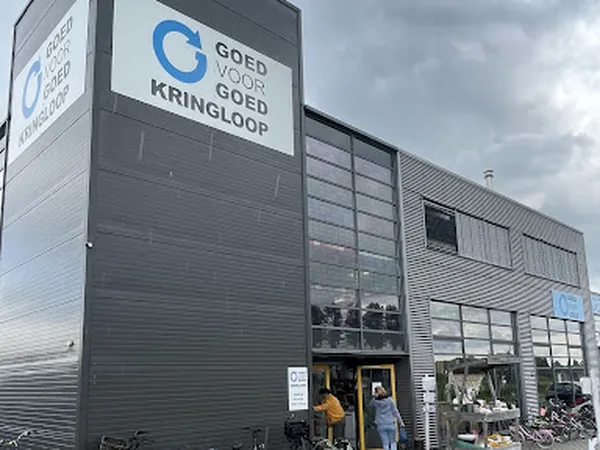 Grijze gevel van kringloopwinkel met blauw logo 'Goed voor Goed Kringloop' en grote ramen, klanten bij ingang