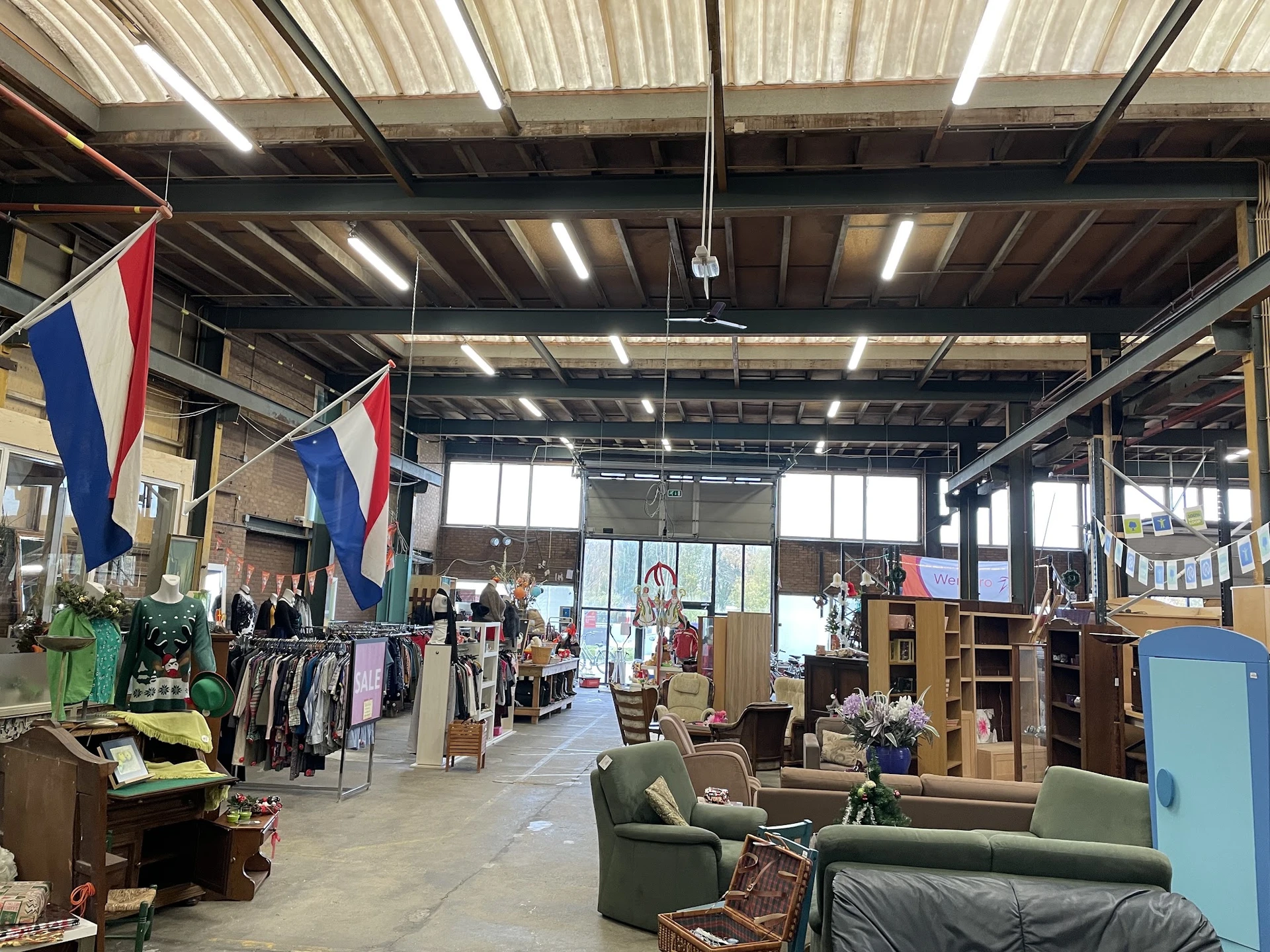 Ruim magazijnachtig interieur van kringloopwinkel met meubels, kleding en diverse spullen, Nederlandse vlaggen, goed verlicht