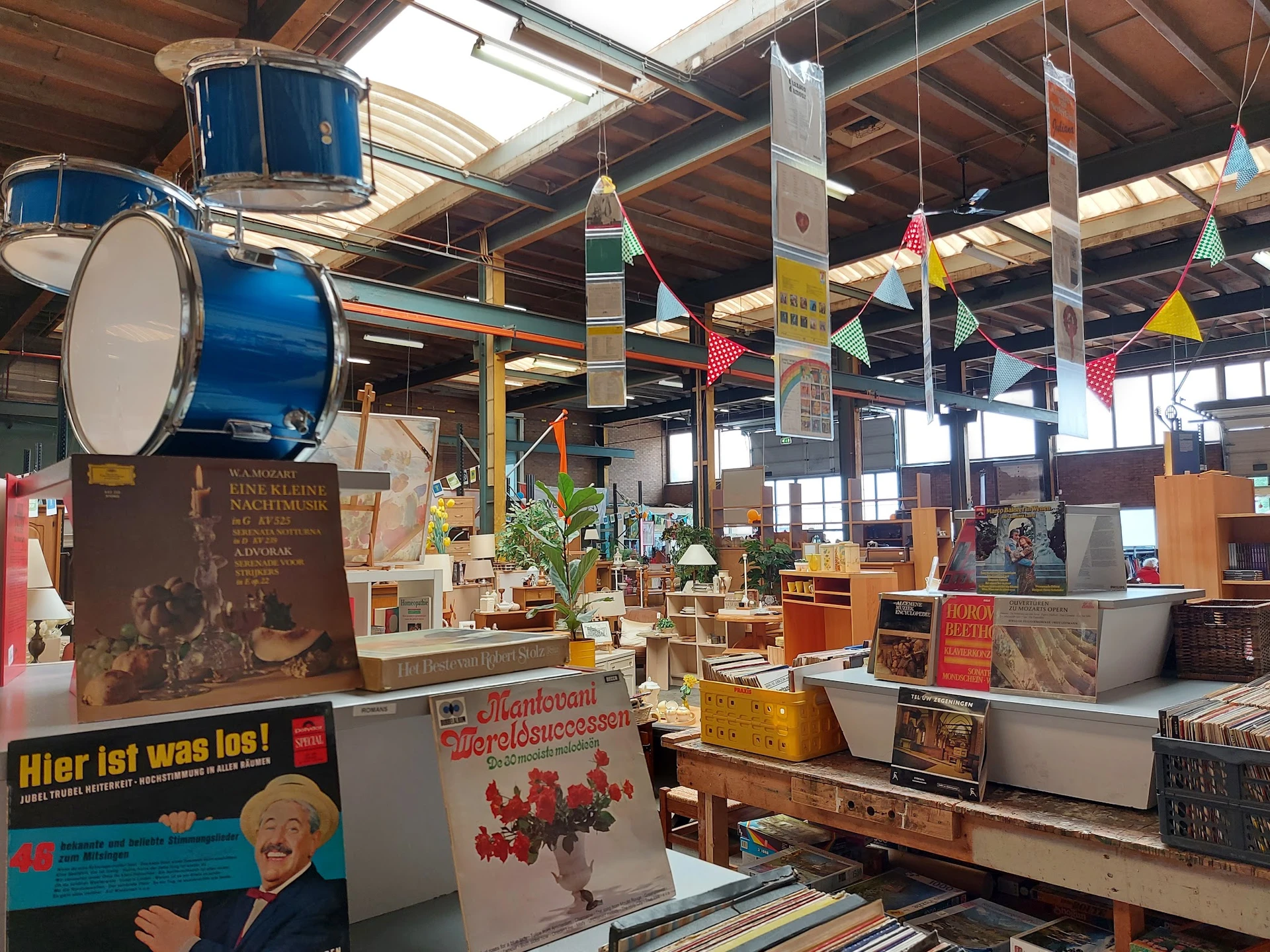 Ruim magazijn met houten balken, hangende blauwe trommels, diverse meubels en spullen op tafels, kleurrijke decoraties aan he