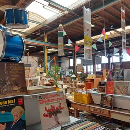 Ruim magazijn met houten balken, hangende blauwe trommels, diverse meubels en spullen op tafels, kleurrijke decoraties aan he