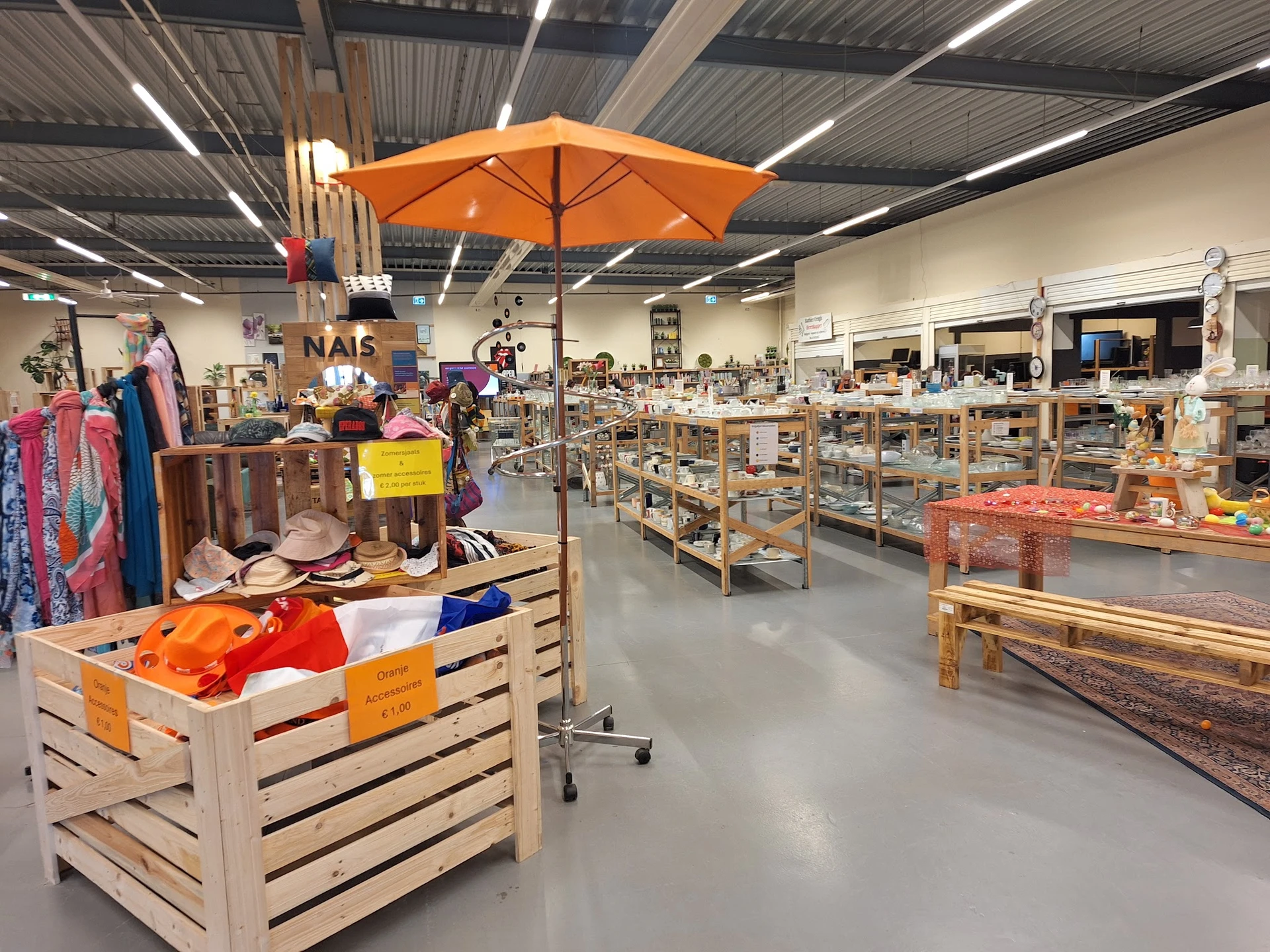 Ruim winkelinterieur met houten stellingen vol spullen, kleding op hangers links, oranje parasol aan plafond