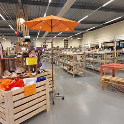 Ruim winkelinterieur met houten stellingen vol spullen, kleding op hangers links, oranje parasol aan plafond