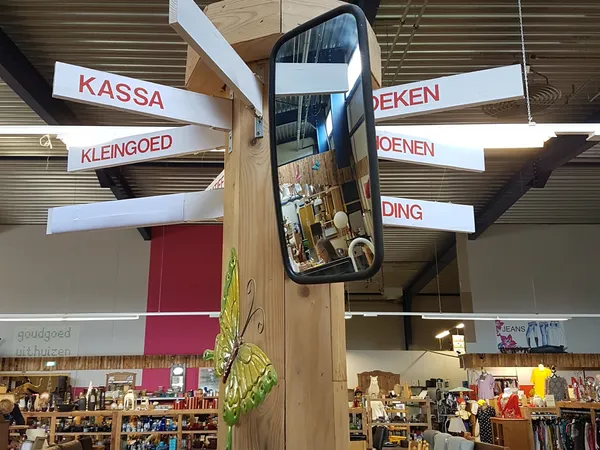 Winkelinterieur met houten pilaar, hangende spiegels en bewegwijzering met Nederlandse teksten zoals 'KASSA', 'KLEINGOED' en 