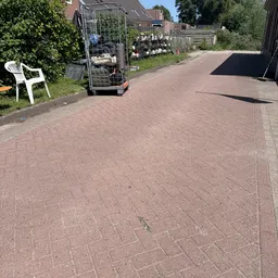 Parkeerterrein met bakstenen bestrating, witte stoel en transportwagen links, schaduw van gebouw rechts