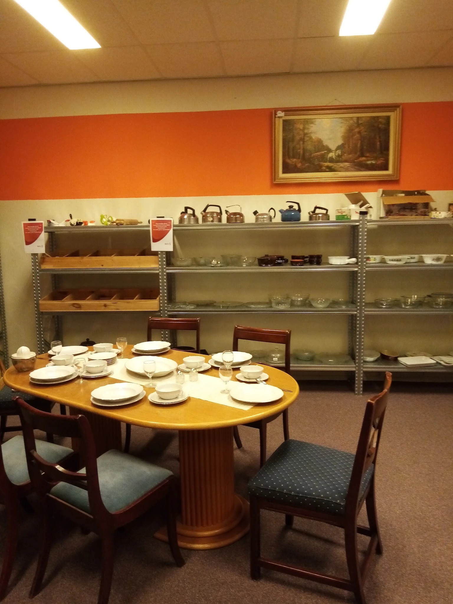 Winkelbinnenkant met serviezen op tafel, stellingen met huishoudelijke spullen en oranje muur