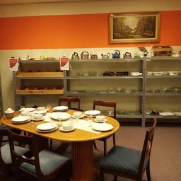 Winkelbinnenkant met serviezen op tafel, stellingen met huishoudelijke spullen en oranje muur