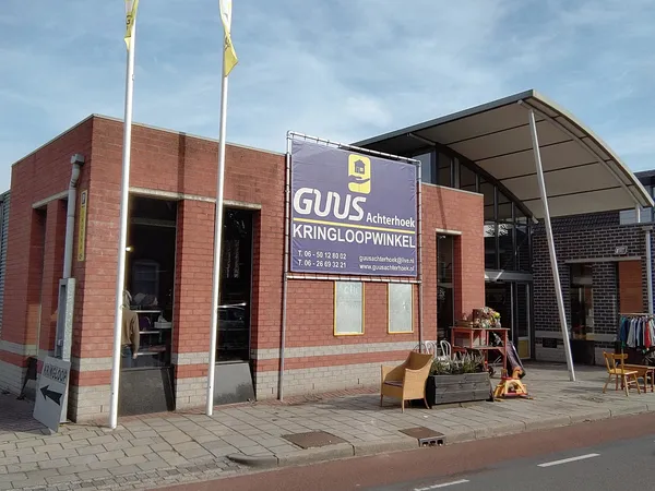 Voorgevel van een rode bakstenen kringloopwinkel met naam- en logo-bord, twee vlaggen, en terras met stoelen