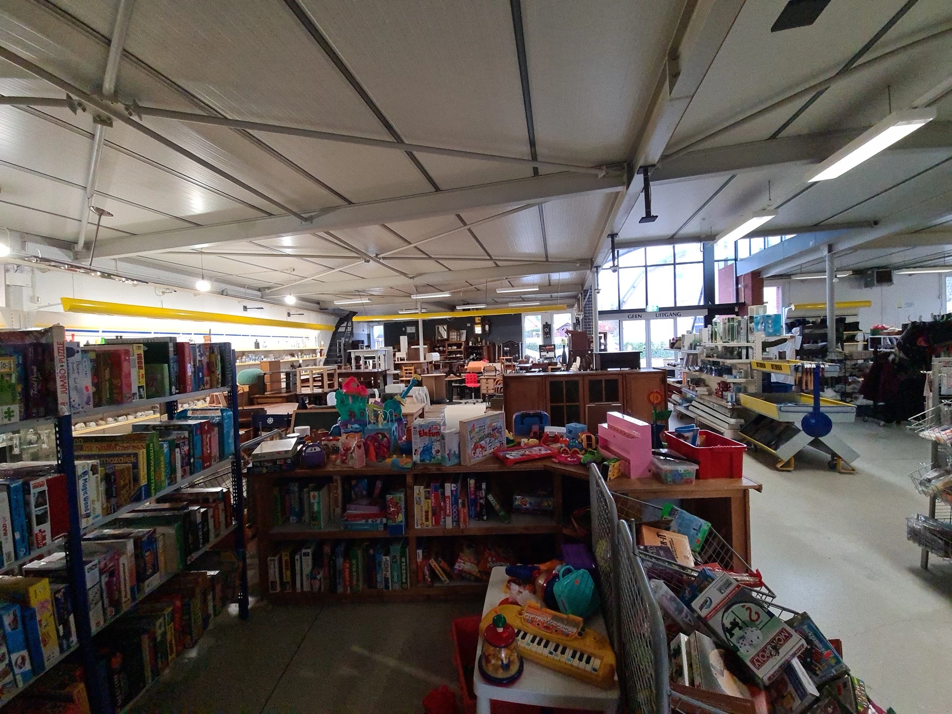 Overzicht van de winkelvloer met boekenrekken, speelgoed, kleding en diverse tweedehands artikelen onder fluorescente verlich