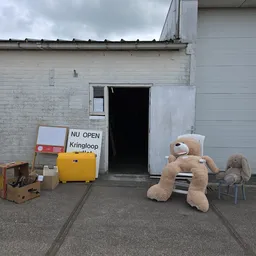 Ingang van kringloopwinkel met uitgestalde spullen: knuffels, koffers, stoelen en diverse tweedehands artikelen
