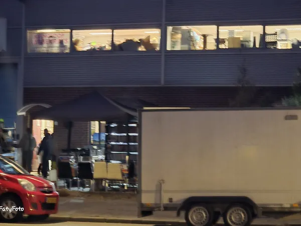 Gevel van kringloopwinkel 's avonds met verlichte ramen en ingang, rode auto en witte aanhanger op straat