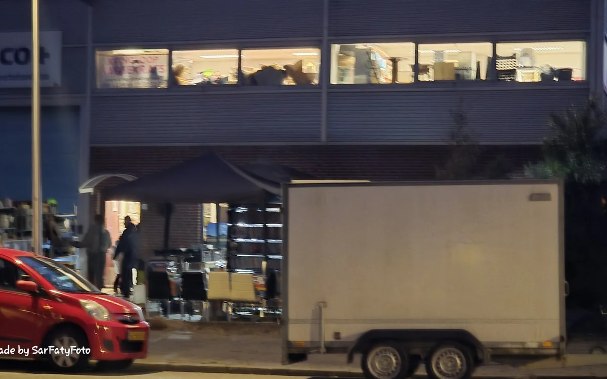 Gevel van kringloopwinkel 's avonds met verlichte ramen en ingang, rode auto en witte aanhanger op straat