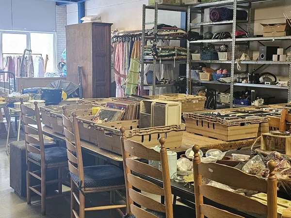 Binnenaanzicht van kringloopwinkel met houten stoelen, dozen, stellingen en kleding op achtergrond