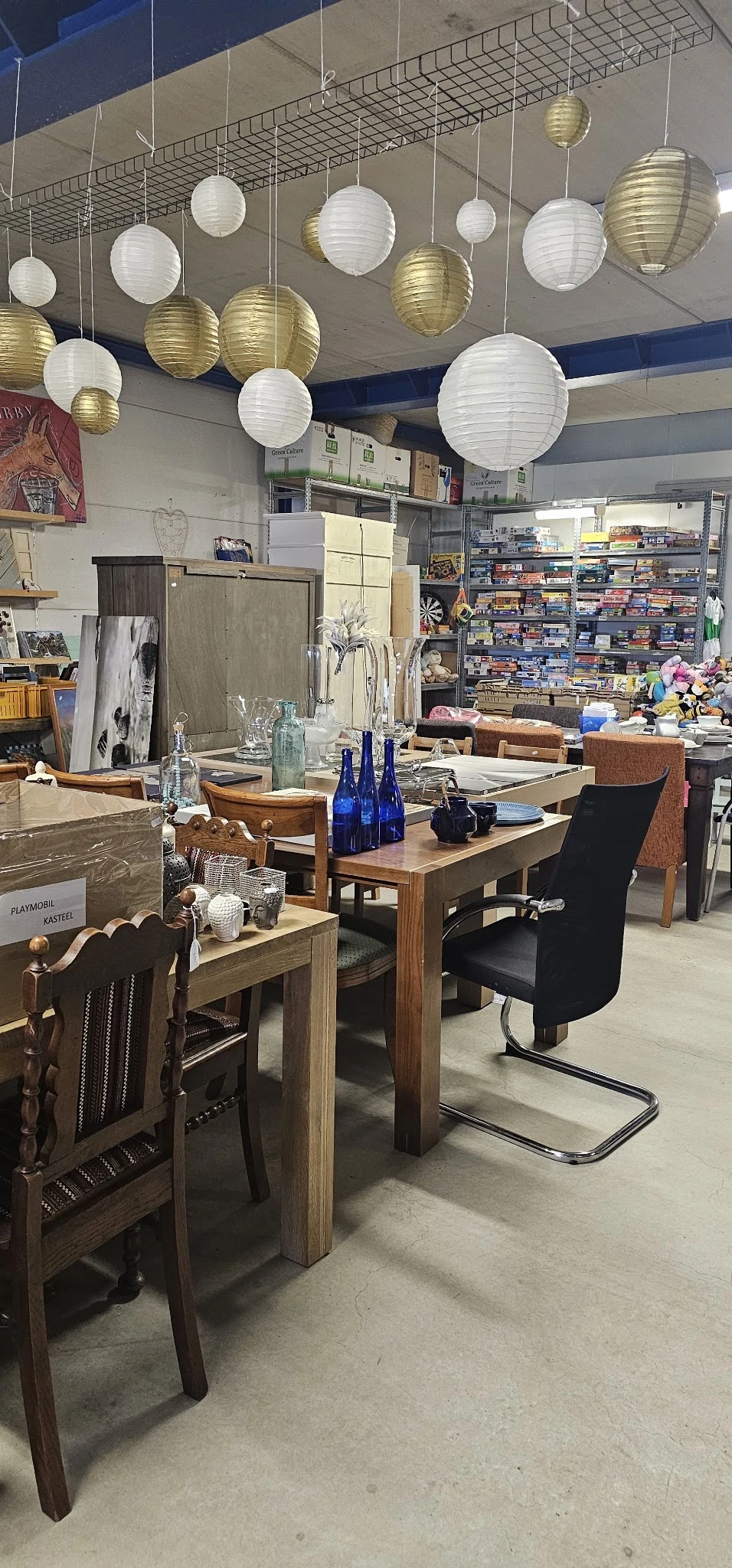 Winkelbinnenruimte met houten tafels, stoelen, blauwe flessen, serviesgoed en schappen vol spullen op de achtergrond