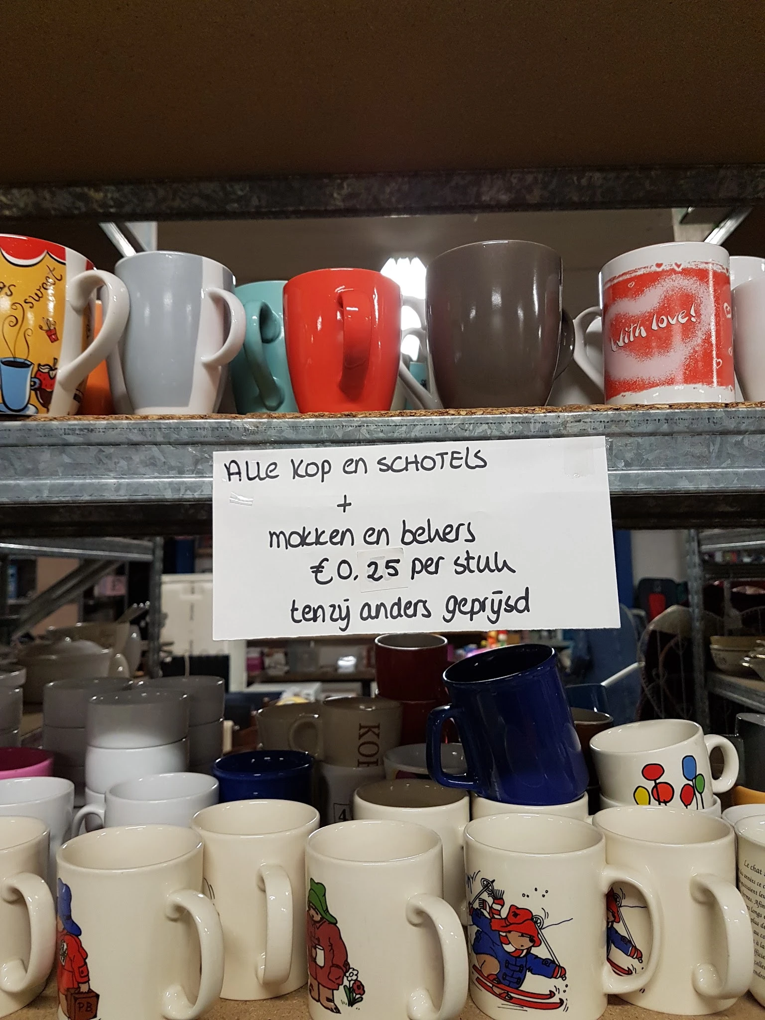 Handgeschreven prijsbord voor koffie en schotels bij een kringloopwinkel met diverse mokken en bekers op de achtergrond