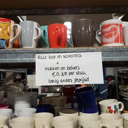 Handgeschreven prijsbord voor koffie en schotels bij een kringloopwinkel met diverse mokken en bekers op de achtergrond