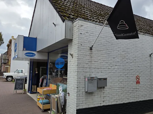 Witte bakstenen gevel van kringloopwinkel met blauwe logo, zwarte vlag met winkellogo, ingang met etalage en gele stellage bu