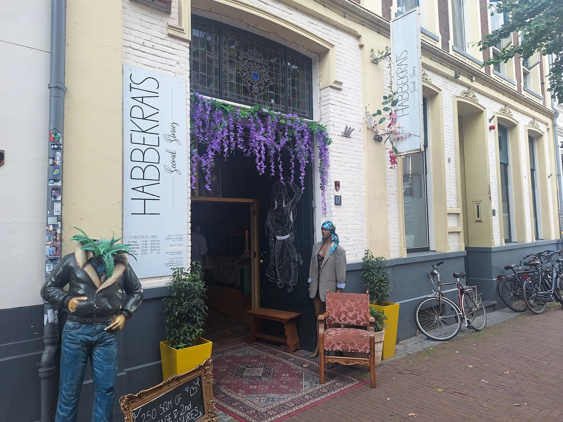 Gevel van kringloopwinkel met witte houten pui, paarse bloemen boven de deur, houten zitbank en gele plantenbakken ervoor