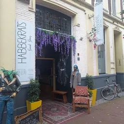 Gevel van kringloopwinkel met witte houten pui, paarse bloemen boven de deur, houten zitbank en gele plantenbakken ervoor