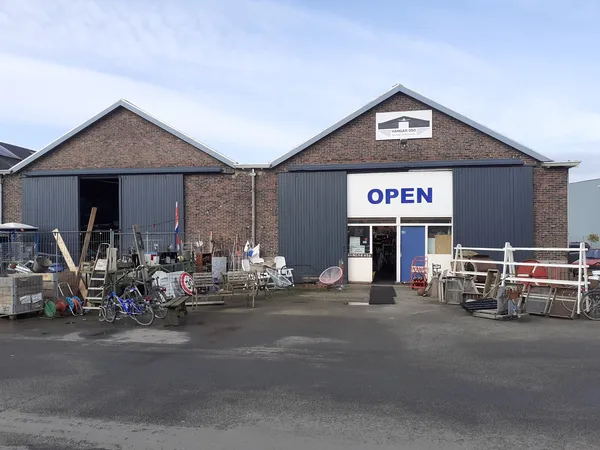 Voorkant van kringloopwinkel met bakstenen gevel, twee units met grijze deuren, OPEN-bord en rommelmarkt-achtig assortiment o