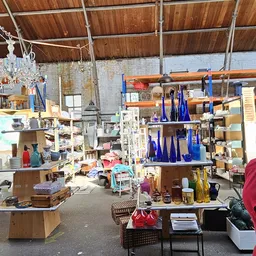 Ruim magazijn met houten balken, stellingen vol decoratieve spullen, blauwe glazen flessen, houten meubels en diverse tweedeh