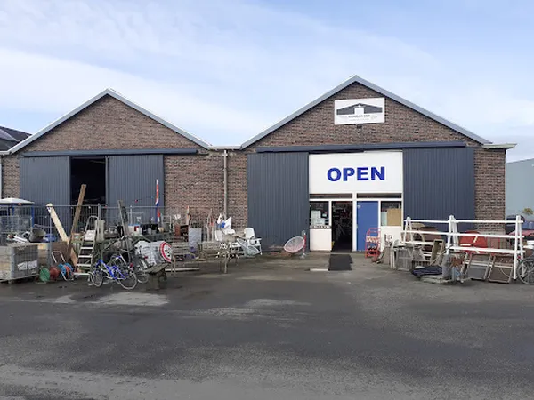 Voorkant van kringloopwinkel met bakstenen gevel, donkergrijs metalen deuren, OPEN-bord en uitgestalde spullen op het terrein