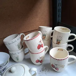 Verzameling witte porseleinen mokken en kopjes met florale en groente-motieven, op een witte plank uitgestald