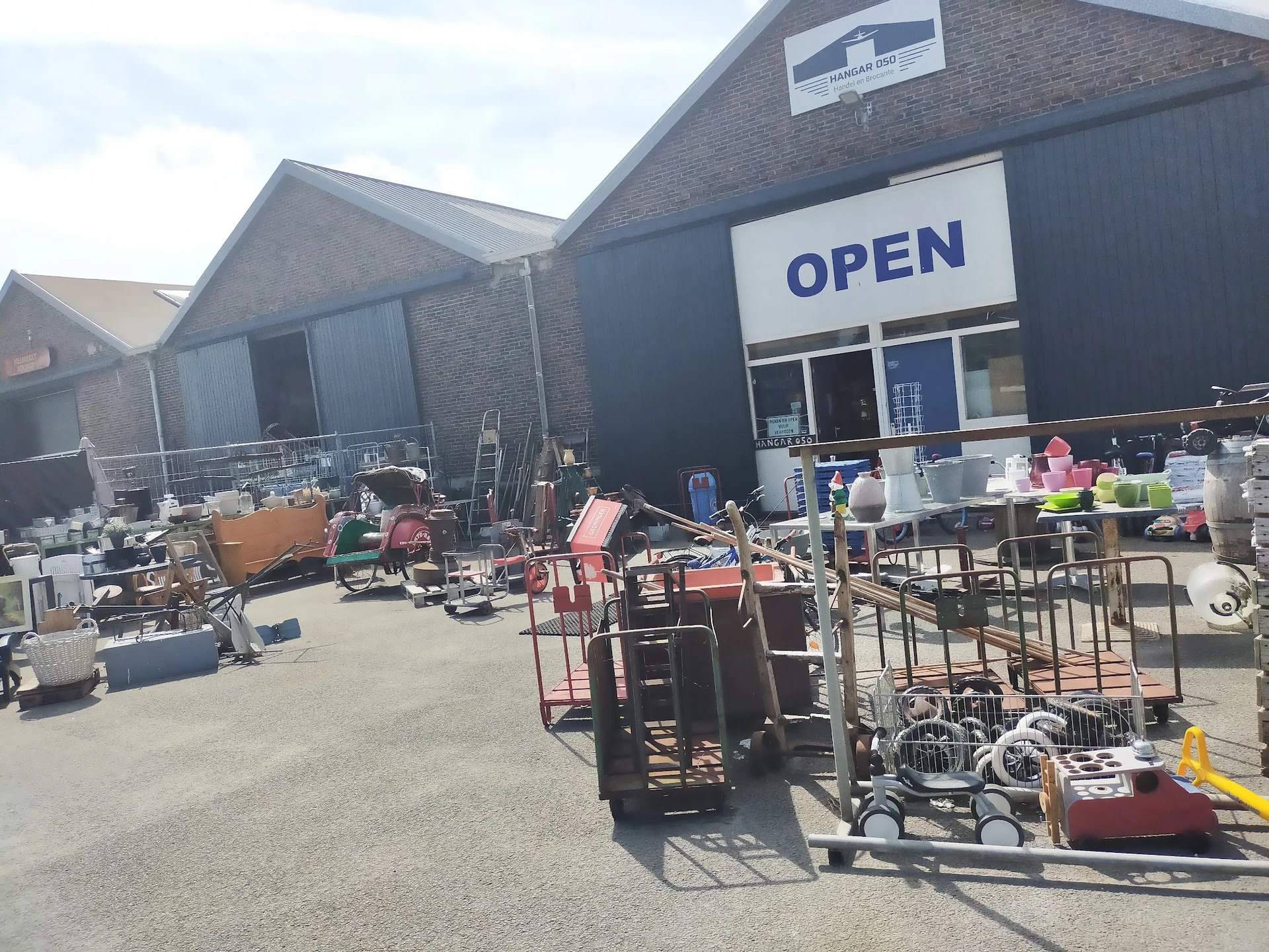 Voorgevel van kringloopwinkel met OPEN-bord, parkeerterrein met gebruikte meubels en spullen