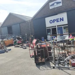 Voorgevel van kringloopwinkel met OPEN-bord, parkeerterrein met gebruikte meubels en spullen