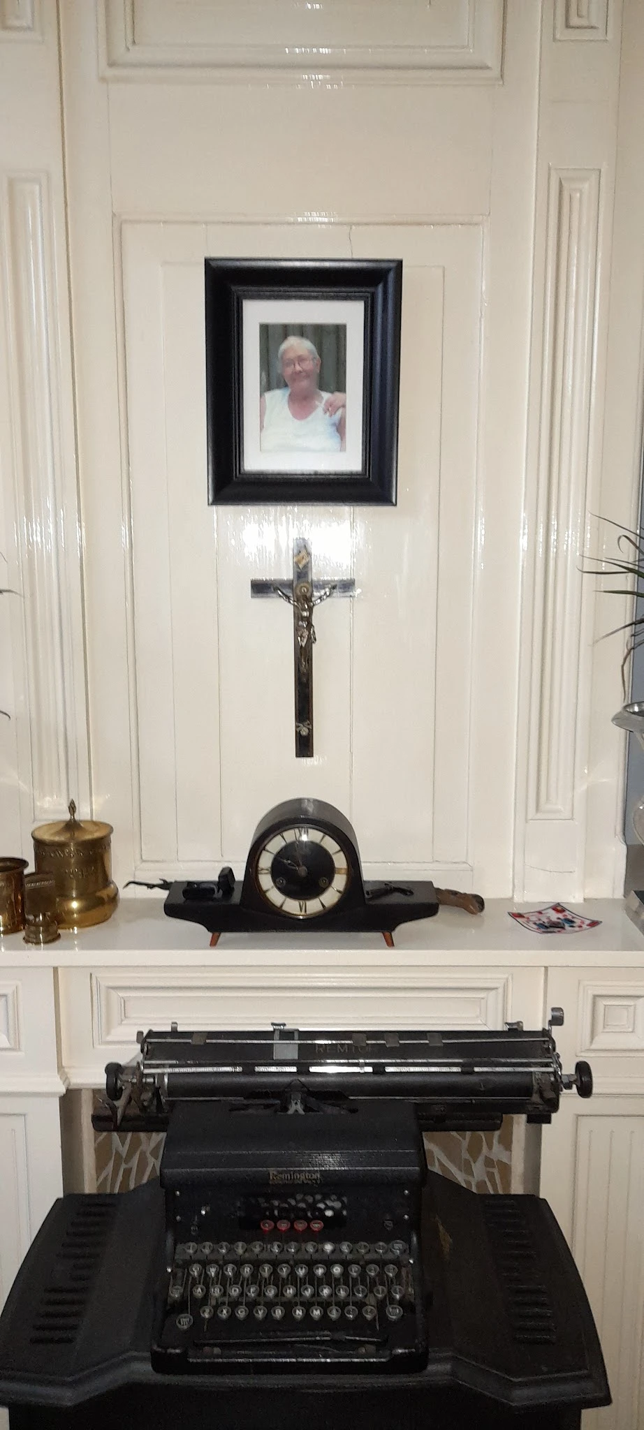 Zwarte vintage tafelklok met witte wijzerplaat en houten voetjes, onder een crucifix aan de muur