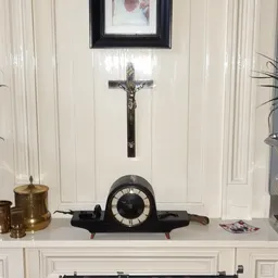 Zwarte vintage tafelklok met witte wijzerplaat en houten voetjes, onder een crucifix aan de muur